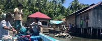 Ost-Indonesien: Drei Männer auf Boot neben Holzhaus auf Stelzen